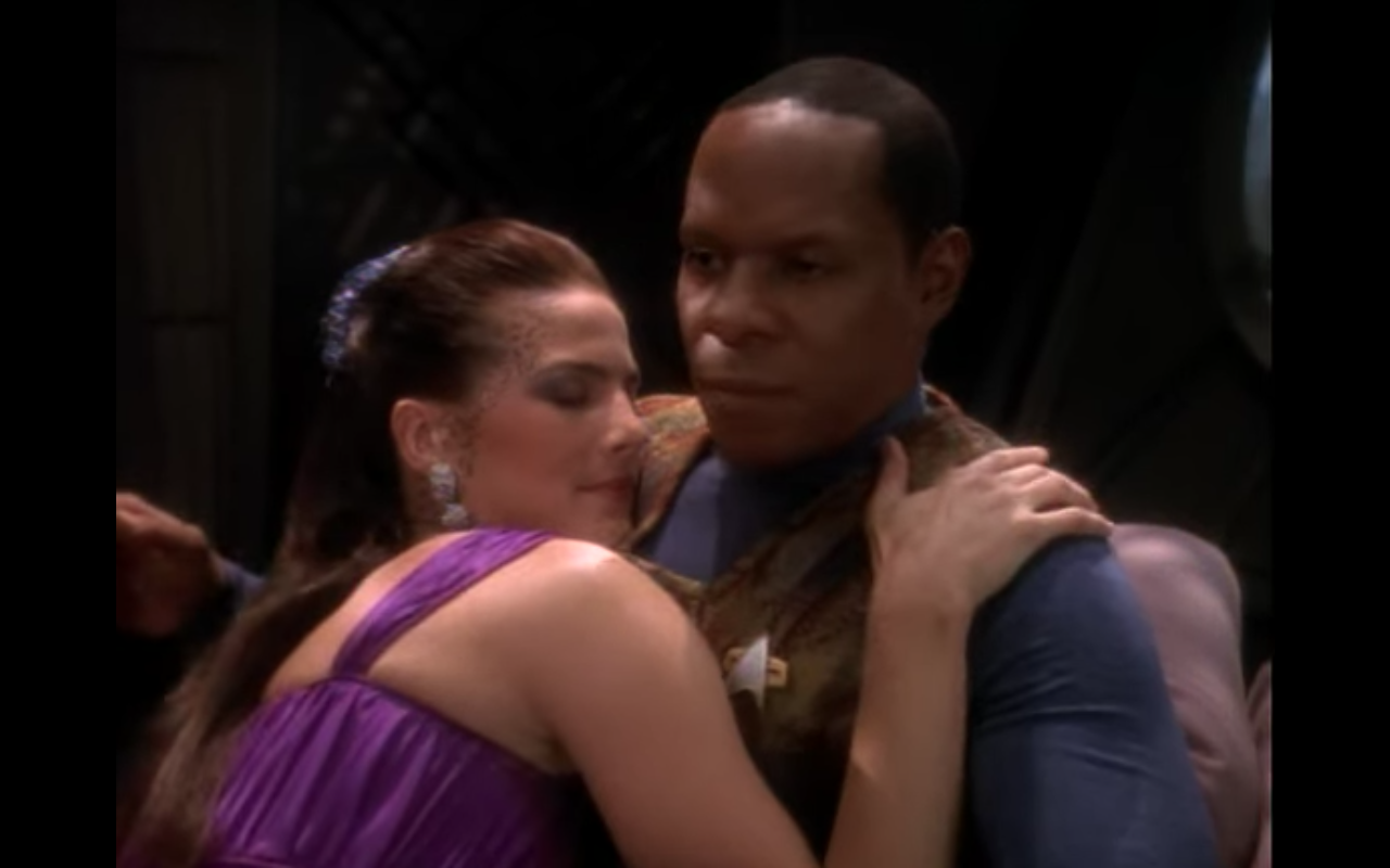 Jadzia in a purple dress, snuggling with an uncomfortable Sisko