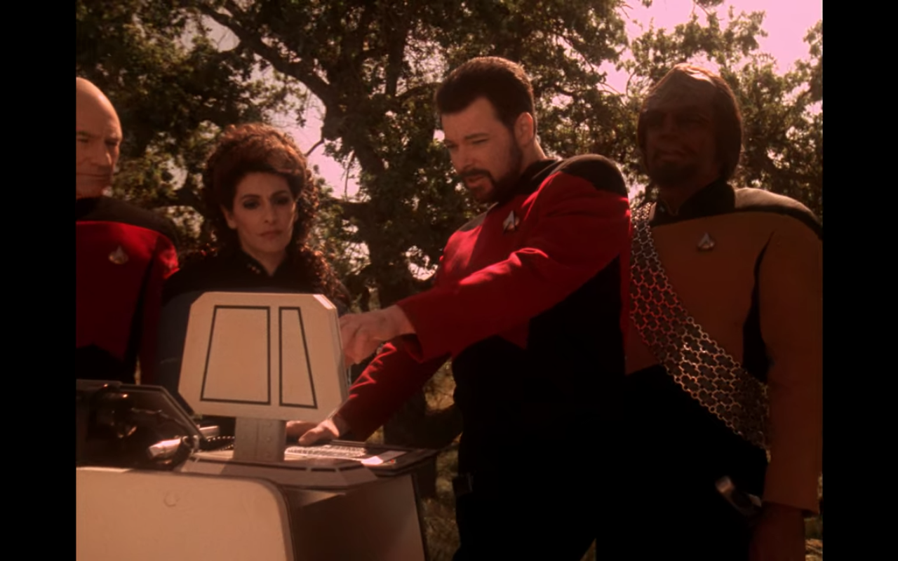 Picard, Troi, Riker, and Worf outdoors looking at a small monitor