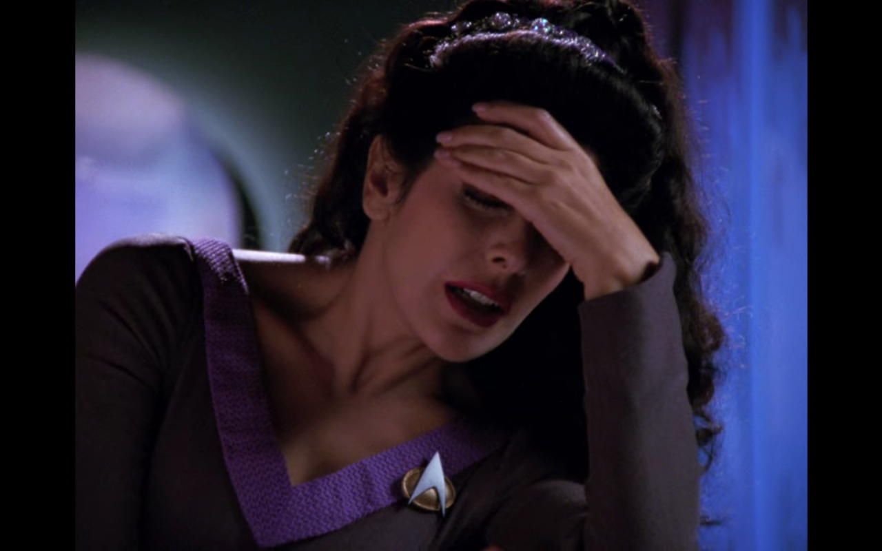 Troi in her purple outfit, holding her hand to her head in pain
