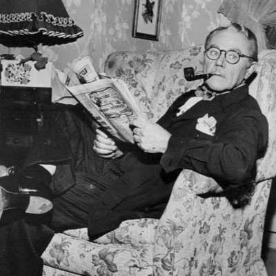 a white man sitting down, reading a newspaper, with a pipe